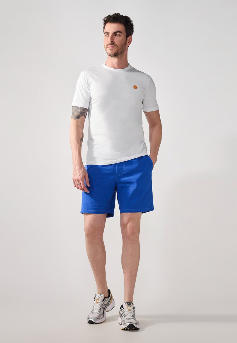 White short-sleeve t-shirt with a small orange emblem, paired with bright blue shorts. Gray and white athletic shoes complete the outfit.