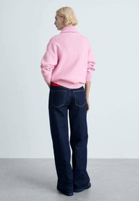 Rosa gestrickter Pullover mit hohem Kragen, kombiniert mit weit geschnittenen, dunkelblauen Jeans. Das Outfit hat eine entspannte Passform und sichtbare Nähte.