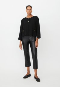 Zwarte cropped jas met gouden knopen, twee steekzakken aan de voorkant en driekwart mouwen, gecombineerd met zwarte faux leren broek en platte schoenen.