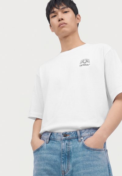Jeune homme portant un t-shirt blanc de la marque Off-White et un jean bleu, les mains dans les poches, debout devant un fond clair uni.