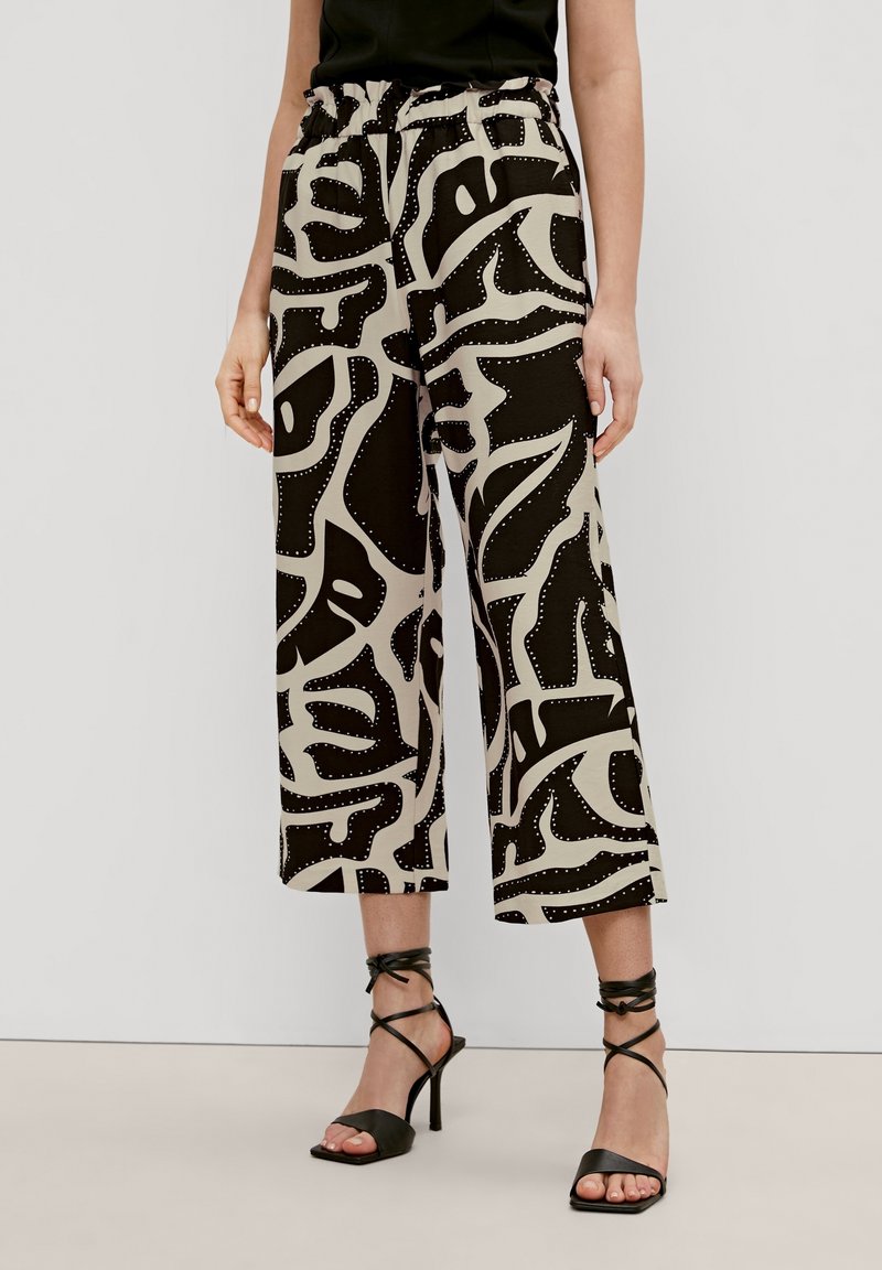 Femme portant un pantalon beige à jambes larges avec des motifs abstraits noirs et des sandales noires à talons hauts avec des brides à la cheville.