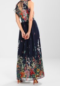 Kvinna klädd i en ärmlös mörkblå maxi-klänning med färgglad blommig tryck och öppen rygg som knyts med en rosett, stående med händerna sammanslagna bakom.