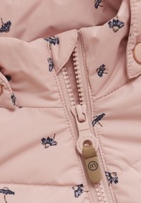 Veste matelassée rose clair avec des motifs floraux navy. Fermeture éclair centrale avec une tirette beige et un détail de logo. Texture de tissu douce et lisse.