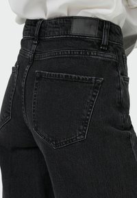 Čierne rifle s vysokým pásom, zadnými vreckami a nášivkou JDY denim, nosené s bielou košeľou zastrčenou dnu.