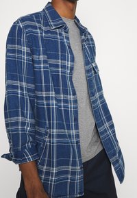 Modrá kostkovaná flanelová košile s bílými pruhy a zapínáním na knoflíky, doplněná šedým tričkem. Má vyhrnovací rukávy a přední kapsy.