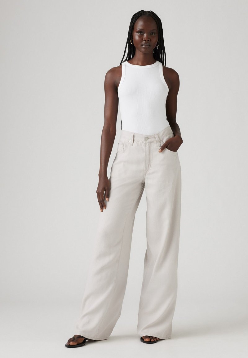 Jeans a gamba larga, di un beige chiaro con vita alta e tasche frontali, abbinati a un top fitted bianco, indossati con sandali neri.