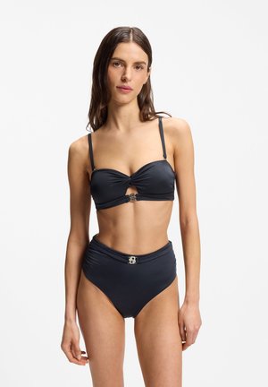 Žena v čiernych bandeau bikinách s kovovými zapínacími detailmi pózuje na bielom jednoduchom pozadí.