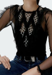 Svart genomskinlig topp med volangärmar, prydd med ett diamantmönster av pärlor. Kombinerad med blå denim, förhöjd med en silverbältesspänne.