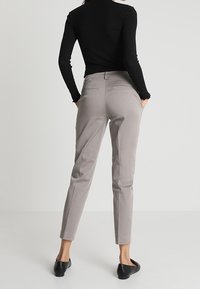 Žena v světlých šedých úzkých kalhotách a černém dlouhém tričku, stojící s rukama v kapsách a v černých balerínách na bílém pozadí.