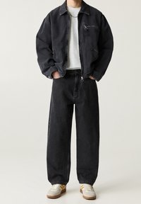 Chaqueta de pana negra con un bolsillo delantero con cremallera, combinada con jeans negros de corte holgado. El modelo lleva una camiseta gris clara y zapatillas casuales.