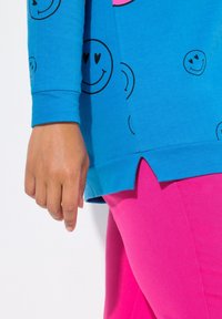 Top blu con stampa di faccina sorridente nera, con spacco laterale. Abbinato a leggings rosa. Mano visibile appoggiata sul capo.