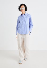 Camicia a righe blu chiaro e bianca con taschino, abbinata a pantaloni larghi in lino beige e sneakers blu chiaro.