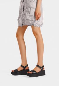 Sandali platform neri con fasce larghe, dettagli con fibbie e suola texturizzata, abbinati a un vestito argento lungo fino al ginocchio con tasche.