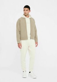 Chaqueta beige claro con cremallera sobre una sudadera con capucha crema, combinada con pantalones jogger crema y zapatillas blancas. Textura suave con diseño minimalista.
