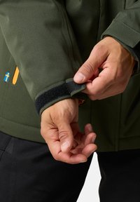 Grön jacka med kardborreband vid manschetterna, visar en hand som justerar remmen. Har en svensk flagga patch och orangea detaljer på tyget.