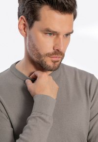 Szary sweter z okrągłym dekoltem wykonany z miękkiego materiału, z ribbowymi ściągaczami przy mankietach i dekolcie. Zbliżenie na dłoń dostosowującą kołnierz.