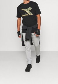 Man som går i svart logot-shirt, ljusgrå joggingsbyxor, svarta sneakers, fingerlösa handskar och en tröja knuten runt midjan.