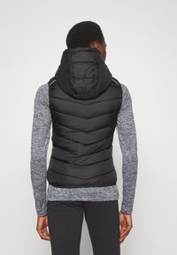 Musta vuorellinen liivi, jossa on huppu ja viistokuvio, valmistettu kiiltävästä materiaalista. Se on puettu harmaan pitkähihasen paidan ja mustien housujen päälle.