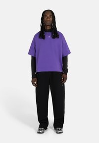 Lilla oversize t-skjorte med korte ermer, båret over en svart langermet topp. Svart løse bukser og metalliske joggesko fullfører antrekket.