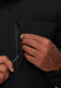Zwarte, met rits gesloten jas van gemêleerd materiaal. Een hand trekt de rits van een borstzakje. Beschikt over verstelbare trekkoorden bij de capuchon.