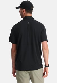 Polo shirt atletico nero con una texture liscia, maniche corte e un logo discreto sulla parte superiore della schiena. Abbinato a pantaloni corti di colore chiaro.