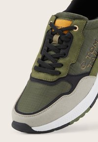 Sneaker verde oliva con materiali in mesh testurizzato e suede, accenti neri e una suola in gomma con motivo. Dettaglio del logo dorato sul lato.