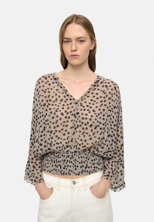 Przezroczysta, beżowa bluzka w czarne grochy, z dekoltem w serek i marszczonym pasem. Długie, miękkie rękawy o lekko luźnym kroju. W połączeniu z białymi jeansami.