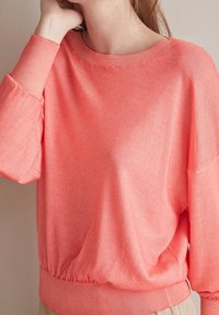 Kobieta w luźnym koralowo-różowym swetrze z długimi rękawami oraz prążkowanymi mankietami, dekoltem i dołem, pozująca z ręką blisko twarzy.