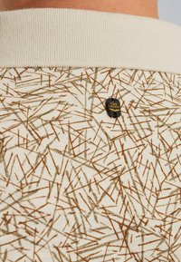 Polo shirt di colore crema chiaro con un motivo brunito testurizzato di linee sparse. Collo a coste e piccolo logo nero accentato sulla parte superiore della schiena.