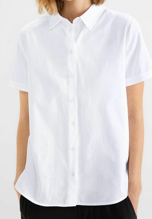 Personne portant une chemise blanche unie à manches courtes avec col, mains partiellement dans les poches d'un pantalon noir, sur un fond uni.
