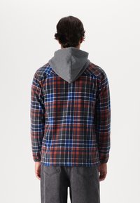 Plaid flannel shirt with red, blue, and black checks, featuring a gray hoodie. Long sleeves, soft texture, and relaxed fit.