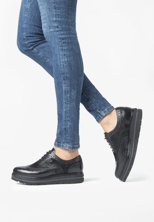 Scarpe stringate nere in pelle con dettagli testurizzati, cuciture rialzate e suola piatta in gomma, abbinate a jeans aderenti blu.