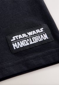 Parche de tela negra con el texto "STAR WARS THE MANDALORIAN" en blanco, cosido al borde de una prenda. Textura lisa, forma rectangular.