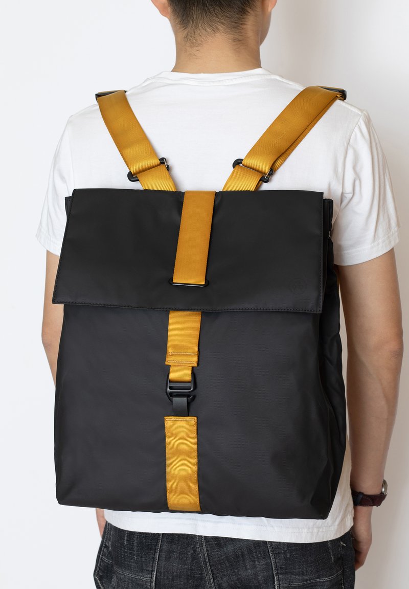 Zaino nero con spalline gialle e chiusura a fibbia. Presenta un coperchio superiore e una texture liscia, sospeso sulla schiena di una persona che indossa una camicia bianca.