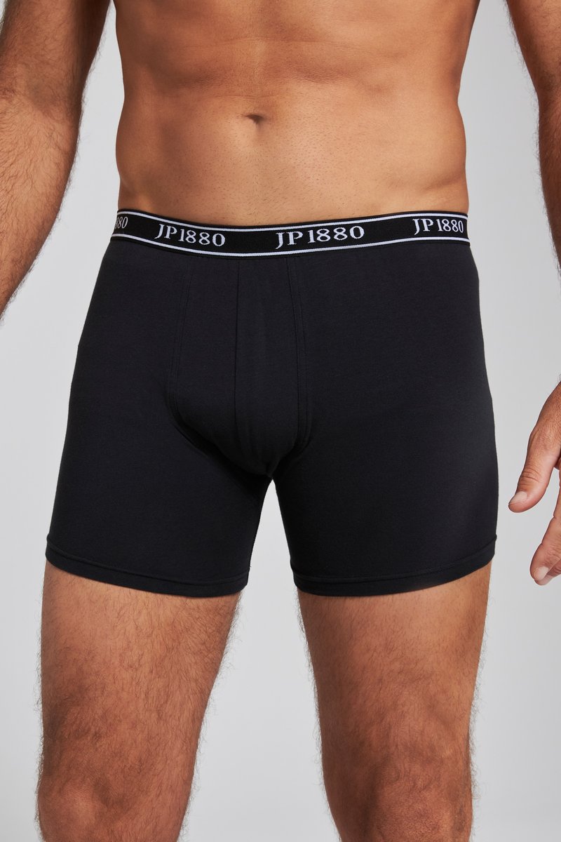 Männlicher Torso, der schwarze Boxershorts mit "JP1880" am Bund trägt, vor einem einfarbig hellgrauen Hintergrund stehend.