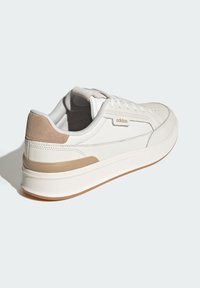 Zapatilla de cuero blanca con acento de ante beige, forma redondeada, suela plana y puntera perforada. Presenta un branding minimalista y cierre con cordones.