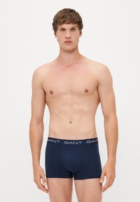 Navy blue boxer shorts with a fitted design and a contrasting white waistband featuring the brand name "GANT" printed repetitively.