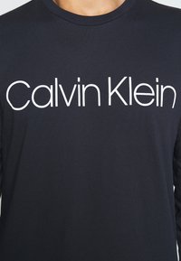 Granatowa bawełniana koszulka z długim rękawem z dużym białym logo "Calvin Klein" na piersi, ribbowanym okrągłym dekoltem i gładką fakturą.