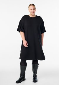 Čierne voľné šaty s krátkymi rukávmi, hladká tkanina, po kolená, skombinované s vysokými čiernymi koženými čižmami s prackami a zipsami.