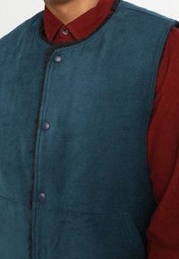 Person in einem türkisfarbenen Cordwesten mit schwarzem Besatz und Druckknöpfen über einem roten Hemd mit Kragen.