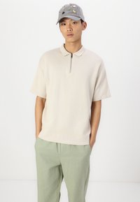 Maglietta polo in maglia beige con colletto a mezza zip abbinata a pantaloni verde chiaro. Il cappellino presenta vari patch colorati e ricami.