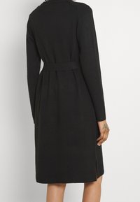 Černé pletené šaty s dlouhými rukávy, páskem v pase, bočními rozparky a žebrovanými manžetami a lemem. Hladká textura a jednoduchý design.