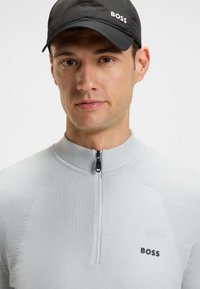 Felpa grigia chiara con zip, texture a coste, colletto alto e logo "BOSS" nero sul petto. Cappellino nero con logo bianco.