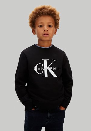 Młody chłopiec z kręconymi włosami, ubrany w czarną bluzę Calvin Klein i ciemne spodnie, stoi z dłońmi w kieszeniach na jednolitym tle.