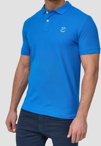 T-shirt polo bleu en tissu lisse. Dispose d'une patte de boutonnage à trois boutons, de manches courtes et d'un petit logo sur la poitrine gauche.