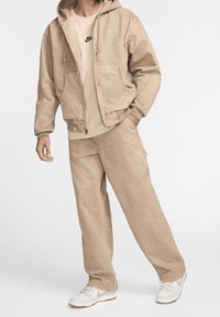 Man i beige Nike-träningskläder med zip-up hoodie, lösa byxor och vita sneakers, stående med ena handen i fickan mot vit bakgrund.