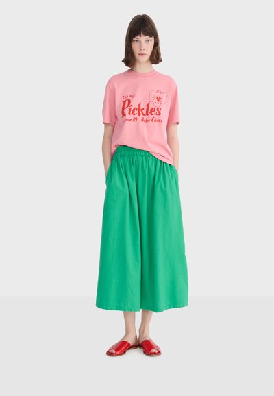 Mladá žena v ružovom tričku s potlačou, širokej zelenej midi sukni a červených otvorených šľapkách stojaca s rukami vreckách pred jednoduchým pozadím.