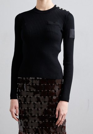 Schwarzes geripptes Langarmshirt mit kleiner Brusttasche, verzierten Schulterknöpfen und strukturiertem Stoff. Getragen mit einem glänzenden dunklen Rock.