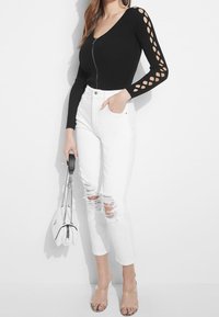 Zwarte geribbelde bodysuit met een rits aan de voorkant en een uitsnijding in het mouwontwerp, gecombineerd met witte versleten jeans en doorzichtige hakken. Witte handtas inbegrepen.
