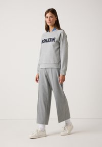 Junge Frau trägt einen grauen Hoodie mit "BONJOUR", graue weit geschnittene Hosen, weiße Socken und weiße Turnschuhe und steht vor einem schlichten Hintergrund.